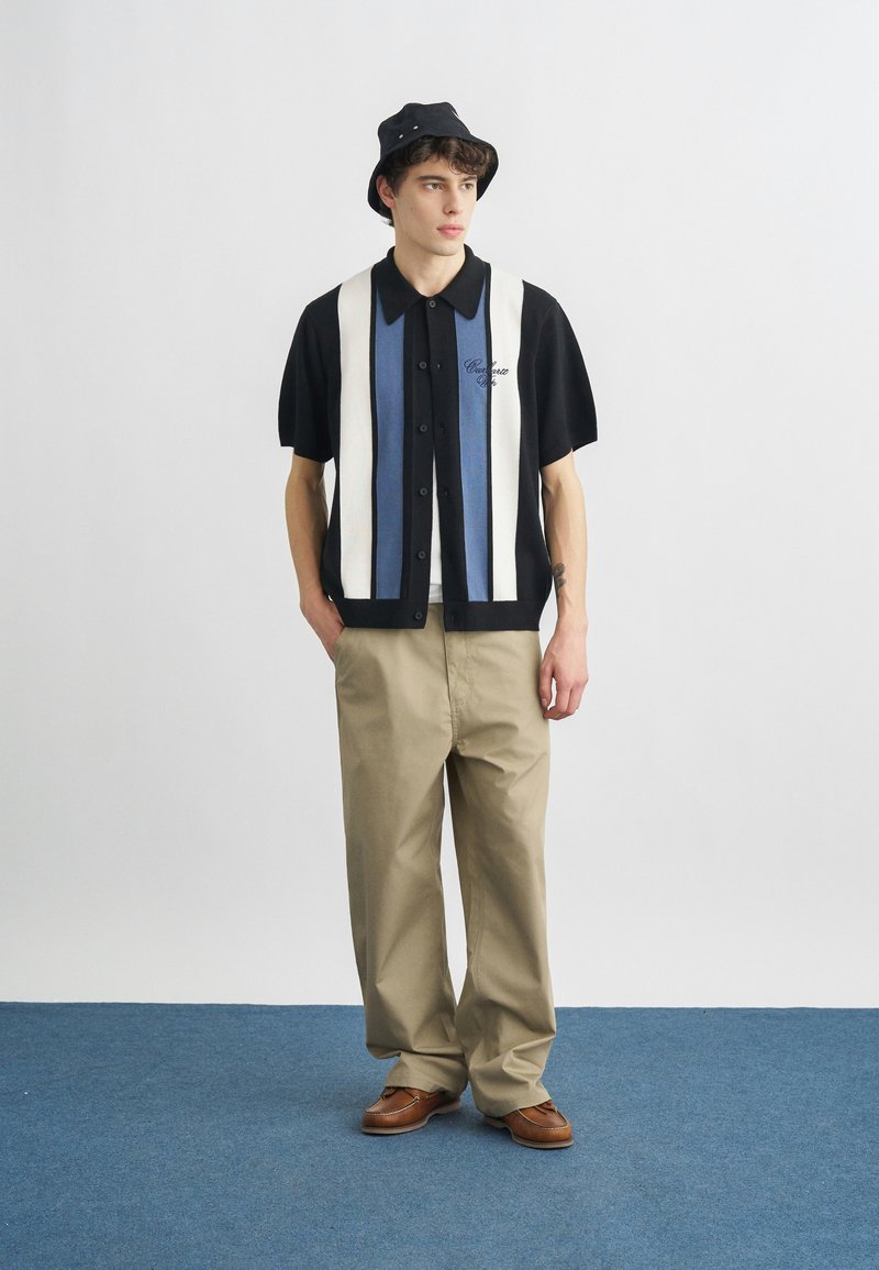 Ung mand iført en sort bøllehat, kortærmet stribet skjorte med knapper, beige bukser med vide ben og brune sko, stående på blå tæppe.