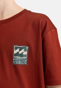 Camiseta de color óxido con mangas cortas, que presenta un parche gráfico cuadrado que representa olas y texto. La tela parece suave y texturada.