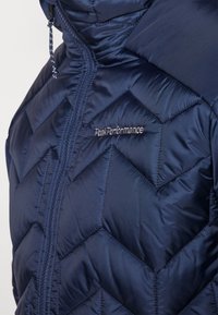 Doudoune bleu marine avec motif de quilting en chevrons, dotée d'une fermeture éclair à l'avant et d'une capuche, marquée "Peak Performance" en broderie argentée.