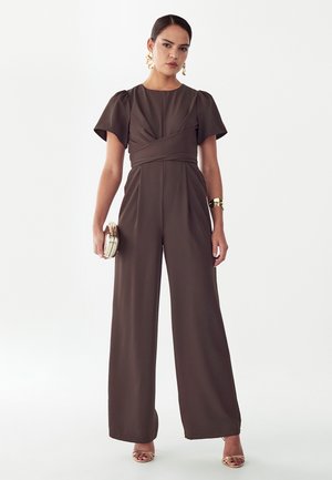 Kobieta w ciemnobrązowym kombinezonie o szerokich nogawkach z krótkimi bufkami, trzyma złotą kopertówkę, nosi złote kolczyki, bransoletkę i buty na obcasie z odkrytymi palcami.