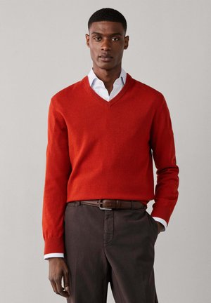 Hombre con suéter naranja de cuello en V sobre camisa blanca, pantalones marrón oscuro y cinturón, de pie frente a un fondo claro y liso.
