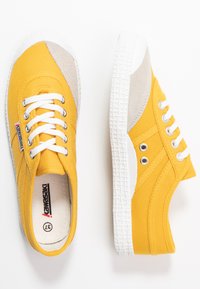 Sneaker in tela gialla con dettagli in suede beige, lacci bianchi e suola in gomma bianca testurizzata. Presenta occhielli sui lati.