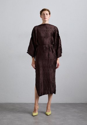 Woman standing against a gray wall wearing a textured, dark brown midi dress with wide sleeves and yellow pointed heels.
