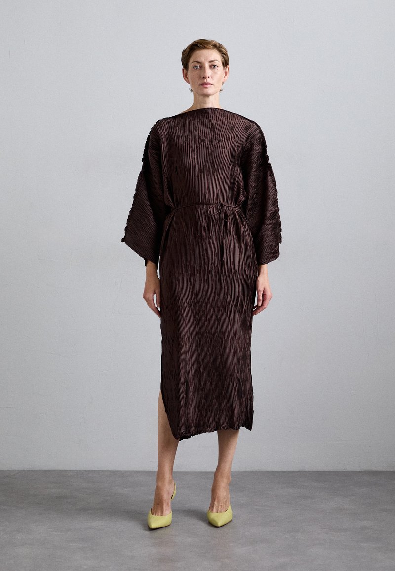 Woman standing against a gray wall wearing a textured, dark brown midi dress with wide sleeves and yellow pointed heels.