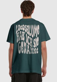 Teal oversized t-shirt med stor hvid tekst på ryggen, der står "Lotosblume hab' ich sie genannt." Blødt bomuldsstof, korte ærmer.