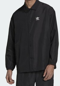 Svart jacka i lättviktsmaterial, med knappstängning, elastiska ärmslut och en broderad vit Adidas-logga på bröstet.