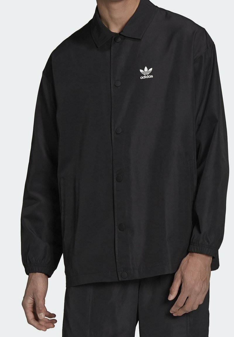 Svart jacka i lättviktsmaterial, med knappstängning, elastiska ärmslut och en broderad vit Adidas-logga på bröstet.