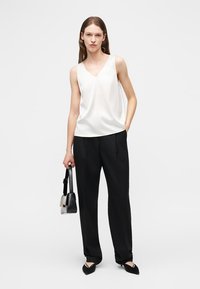 Γυναίκα με λευκό αμάνικο μπλουζάκι, μαύρο παντελόνι φαρδύ, μαύρα μυτερά παπούτσια, κρατώντας μαύρη και λευκή τσάντα, στέκεται μπροστά σε λευκό φόντο.