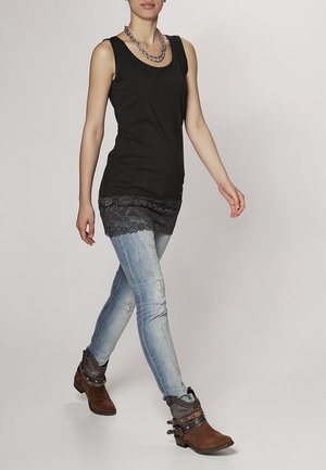 Vrouw loopt in een zwart mouwloos topje met kanten randje, lichtblauwe versleten skinny jeans, bruine enkellaarsjes en gelaagde zilveren kettingen.