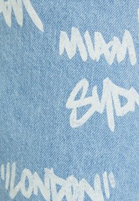 Ljusblå denimtyg med ett vitt graffiti-inspirerat textmönster. Texten innehåller namn på städer i ett tydligt, avslappnat typsnitt.