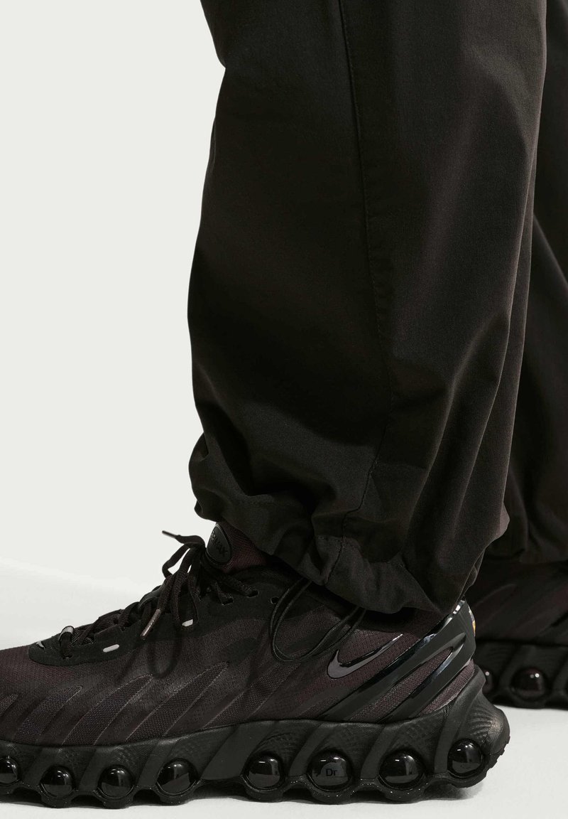 Sneakers nere Nike con suole a bolle trasparenti, indossate con pantaloni neri ampi su uno sfondo chiaro e semplice.