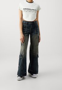 Afgeknot witte t-shirt met zwarte tekst en rand; wijde, vervaagde denim jeans met cargozakken en lichte slijtage; witte sneakers.