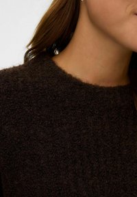 Frau in einem dunkelbraunen strukturierten Strickpullover, Nahaufnahme des Halsausschnitts, des Nackens und eines Teils des Kiefers.