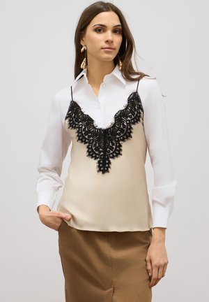 Beige Satin-Top mit schwarzer Spitzenoverlay und verstellbaren Trägern, mit einem V-förmigen Spitzenmuster am Ausschnitt. Lange weiße Hemdsärmel.