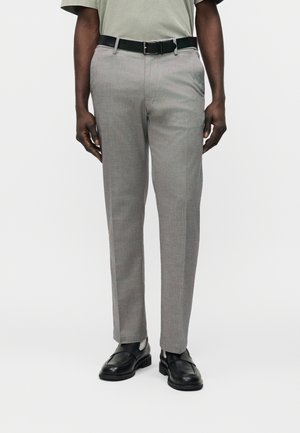 Man draagt lichtgrijze, recht gesneden broek, zwarte leren riem, zwarte loafers en een lichtgekleurde, ingestopte blouse.