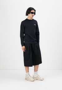Felpa navy con polsini a coste, pantaloni corti neri fino al ginocchio, calze bianche e sneaker crema con suola textured. Occhiali da sole neri presenti.