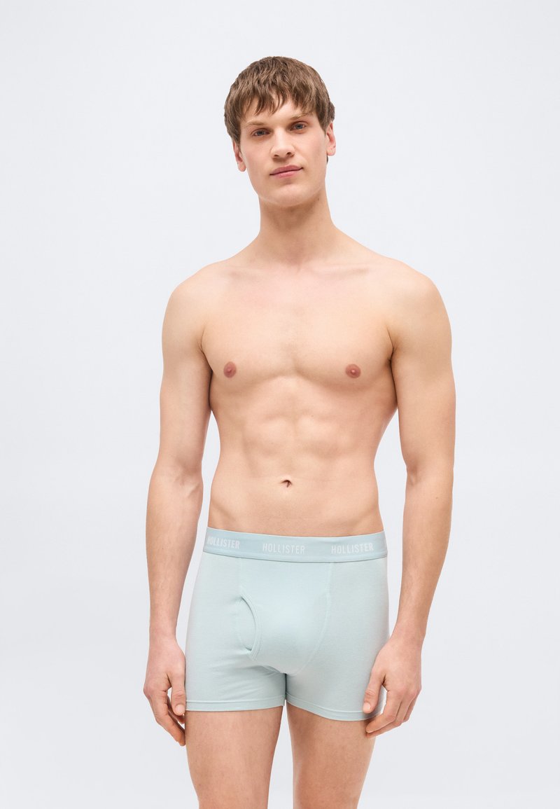 Νέος άνδρας με κοντά καφέ μαλλιά φορώντας ανοιχτό μπλε μπόξερ με λουρί μέσης "Hollister", στέκεται μπροστά σε απλό φόντο.