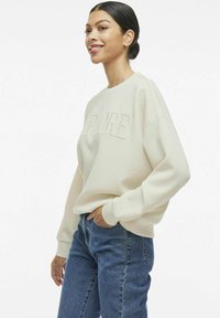 Sudadera crema con letras en relieve "SPORE", corte relajado, puños anchos, combinada con jeans azules, mostrando un estilo casual.