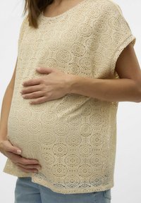 Top de encaje beige con patrón circular, mangas cortas y corte suelto, usado sobre una barriga redondeada. Textura suave con detalle de tejido visible.