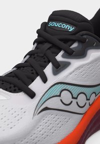Κοντινό πλάνο λευκού παπουτσιού τρεξίματος Saucony με μαύρα κορδόνια, λογότυπο της μάρκας σε τιρκουάζ, υφή υφάσματος και έντονη πορτοκαλί άκρη σολας.