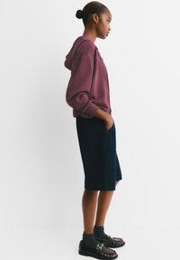 Bordeauxroter Hoodie mit Kängurutasche, kombiniert mit navyblauen Shorts. Schwarze Schuhe mit einer Schnalle und sichtbare graue Socken. Einfaches, lässiges Outfit.