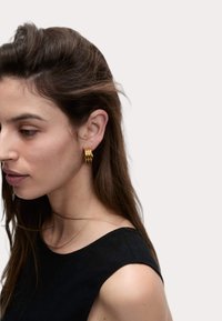 Pendientes de aro dorados con un diseño texturizado, que muestra un patrón acanalado. El modelo tiene el cabello largo y oscuro, y lleva una blusa negra.