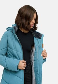 Jasnoniebieska pikowana kurtka z kapturem, z miękkim szarym podszyciem, zapinana na zamek, z bocznymi kieszeniami. Gładka tekstura z detalami szwów.