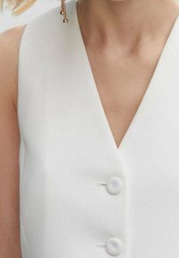 Biała bezrękawnikowa bluzka w serek z dwoma guzikami z przodu, noszona przez osobę z wiszącymi złotymi kolczykami widocznymi z jednej strony.