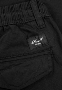 Nahaufnahme von schwarzen elastischen Hosen mit einem genähten Gesäßtasche und einem "Reell est. 1997" Markenetikett.