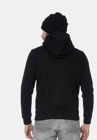 Sudadera negra con capucha, puños y dobladillo acanalados, llevada con un gorro negro. Muestra una textura de tela suave y un diseño ajustado.