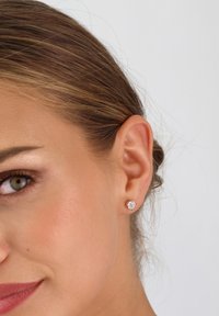 Goldfarbene Ohrstecker mit einem runden, klaren Strassstein in der Mitte, vor einem glatten Hauttextur, mit einer Nahaufnahme des Ohrs.