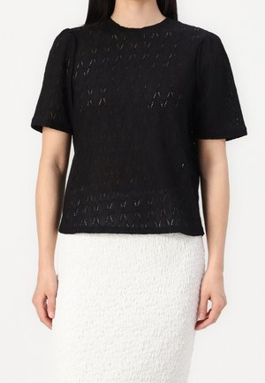 Kobieta ubrana w czarny top z krótkim rękawem i wzorem z koronki, w połączeniu z teksturowaną, białą spódnicą sięgającą kolan, stojąca na jednolitym tle.