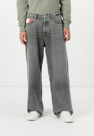 Baggy grå denimjeans med en falmet tekstur, med en rød og hvid lappet lomme detalje. Båret med hvide sneakers, hvilket viser en bred benpasform.