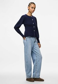 Tumši zils adīts žakets ar apaļu kakla izgriezumu, pogu aizdari un ribotas piedurknes. Saskaņots ar gaiši ziliem brīvi pieguļošiem džinsiem un brūniem apaviem.