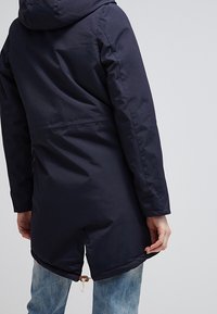 Cappotto imbottito blu navy con cappuccio, dotato di coulisse in vita e orlo posteriore più lungo. Tessuto liscio, dettagli hardware minimali.