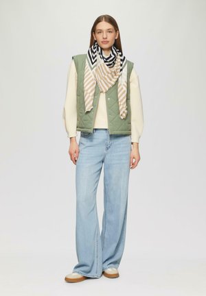 Donna in piedi che indossa un gilet verde trapuntato, un maglione crema, una sciarpa a righe, jeans larghi azzurri chiari e scarpe beige su sfondo uniforme.