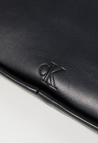 Črna usnjena torba z odtisnjenim logotipom "cK". Gladka tekstura s stilskim oblikovanjem in ukrivljenimi obrobami ob robovih.