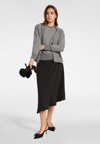 Grau gestrickter Cardigan mit langen Ärmeln über einem grauen Oberteil, kombiniert mit einem schwarzen Midirock und schwarzen Slingback-Absätzen mit einer Schleifenakzent.