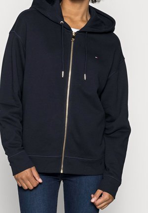 Personne portant un sweat à capuche bleu marine zippé avec un petit logo sur la poitrine et un jean bleu sur un fond uni.