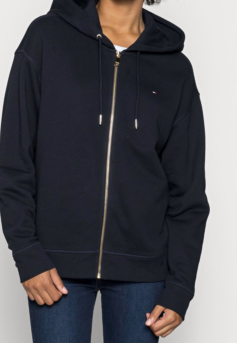 Personne portant un sweat à capuche bleu marine zippé avec un petit logo sur la poitrine et un jean bleu sur un fond uni.