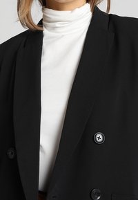 Czarny dwurzędowy żakiet z dużymi guzikami na białym golfie. Żakiet ma strukturalne klapy i gładką teksturę materiału.