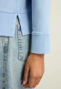 Lyseblå sweatshirt med ribbede manchetter, med et let cropped design og sideslids. Båret over lyse denimjeans.