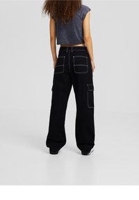 Pantalones cargo negros de pierna ancha, con costuras blancas en contraste, múltiples bolsillos y una cintura alta, combinados con un top gris corto.