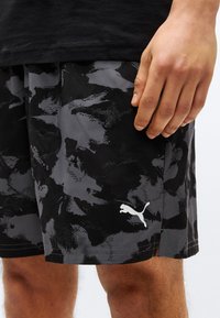 Δεξί χέρι ακουμπισμένο στο πλευρό ενός ατόμου που φοράει μαύρα και γκρι καμουφλαρισμένα σορτς Puma και μαύρο πουκάμισο.