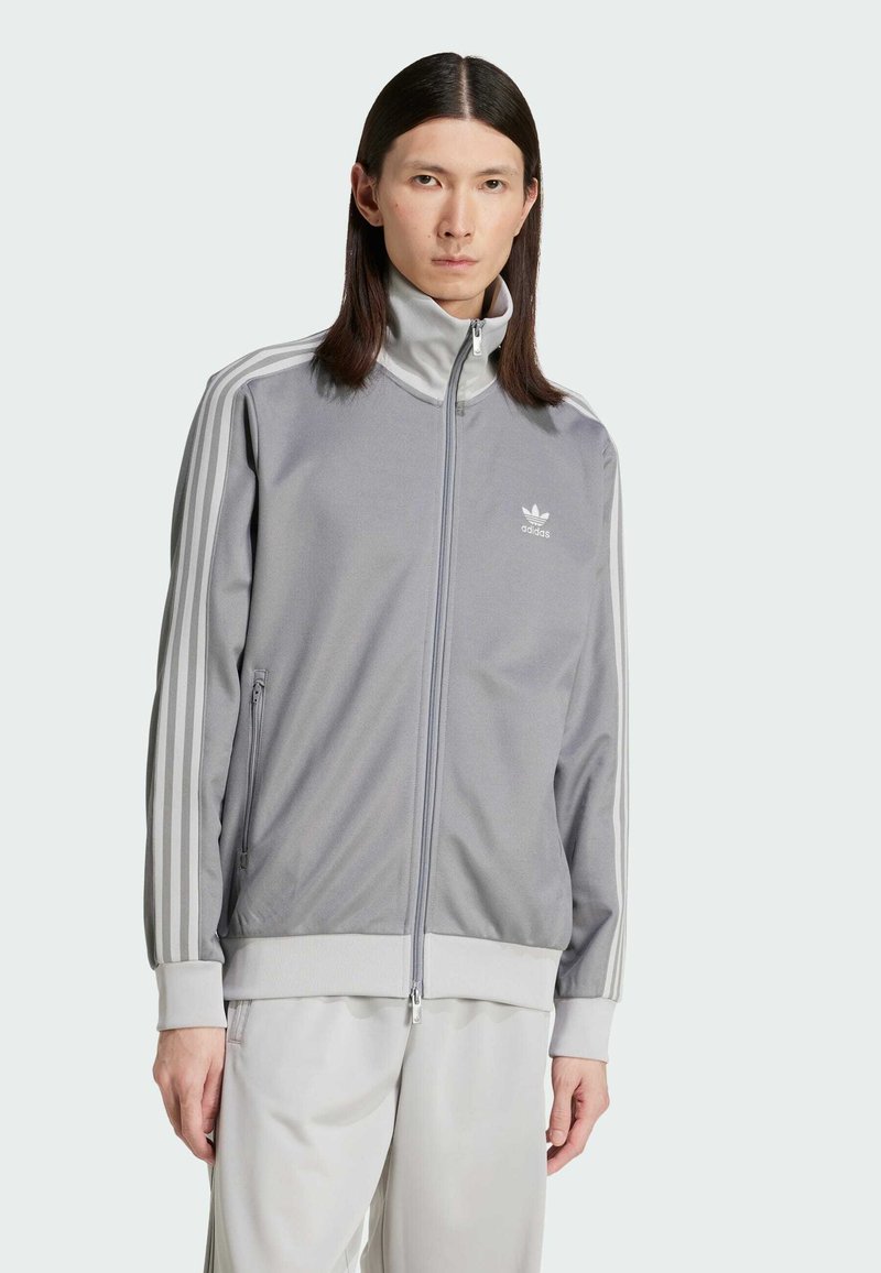 Siva zip-jakna s visokim ovratnikom, bijelim Adidas logotipom i tri bijele pruge duž rukava. Glatka tkanina s opuštenim krojem.