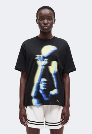 Personne portant un t-shirt noir avec un graphisme bleu et jaune représentant un joueur de basket atteignant un ballon, associé à un short blanc avec des rayures noires.