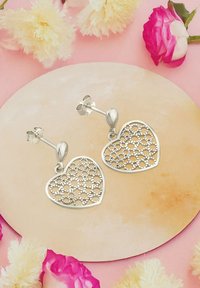 Silverfärgade hjärtformade örhängen med intrikata öppna mönster och en polerad yta, uppvisade på en rund neutral yta.