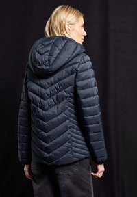 Veste matelassée marine avec un motif en chevrons, design matelassé et capuche. Présente une silhouette ajustée et un tissu texturé.