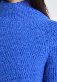 Blauer Strickpullover mit hohem Kragen, der ein geripptes Muster und eine weiche, flauschige Oberfläche aufweist. Nahaufnahme, die die Details des Stoffes betont.