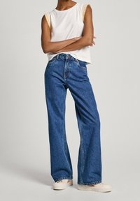 Vit utan ärm topp med scalloped kanter, parat med hög midja blå vida jeans och ljusa avslappnade skor.